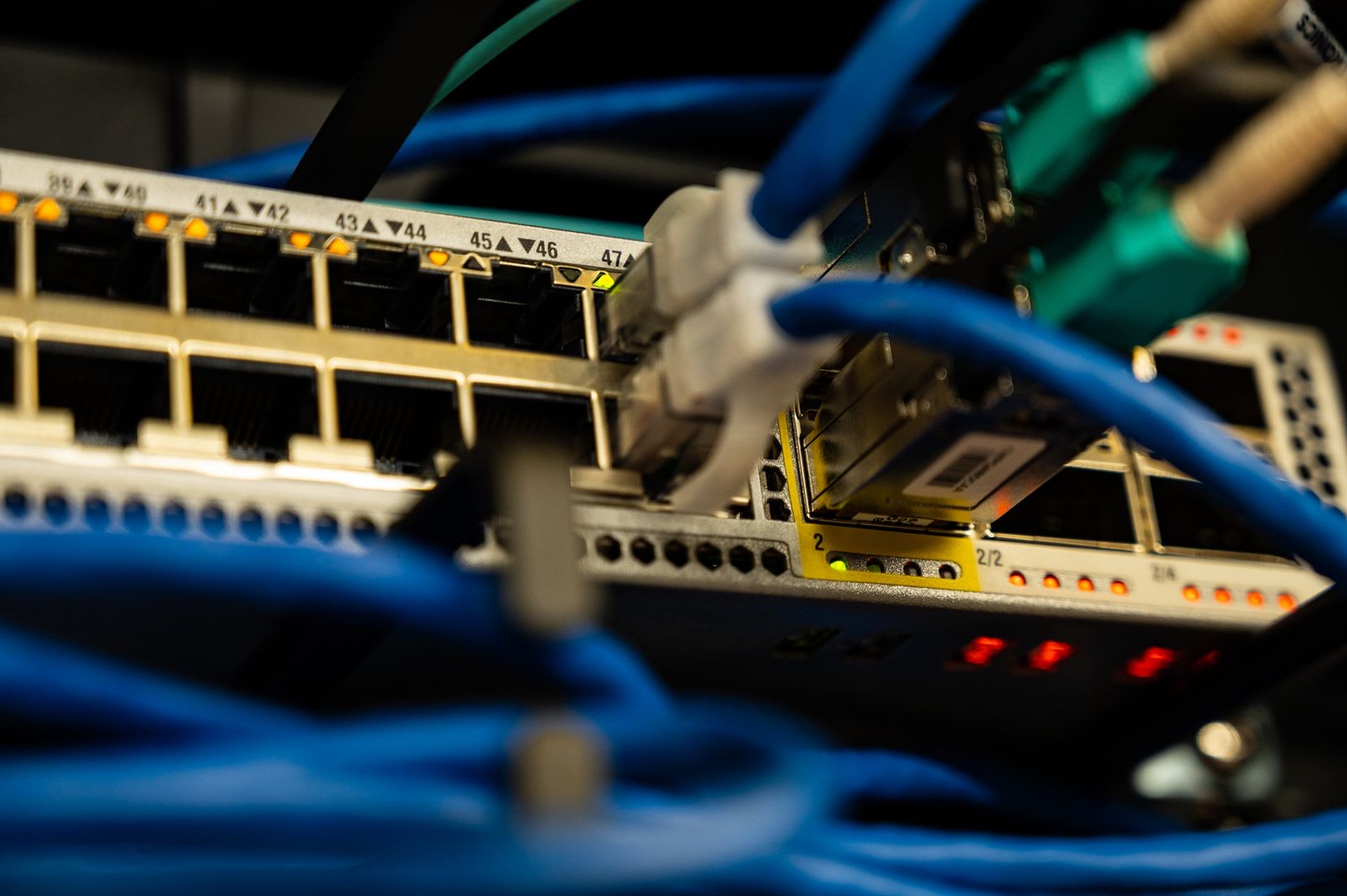 Close-up of the front of a network switch with numbered gold RJ45 ports from 39 to 47, a row of glowing amber link lights, blue patch cables looping across the foreground and a few teal connectors and red status LEDs below, against a dark background