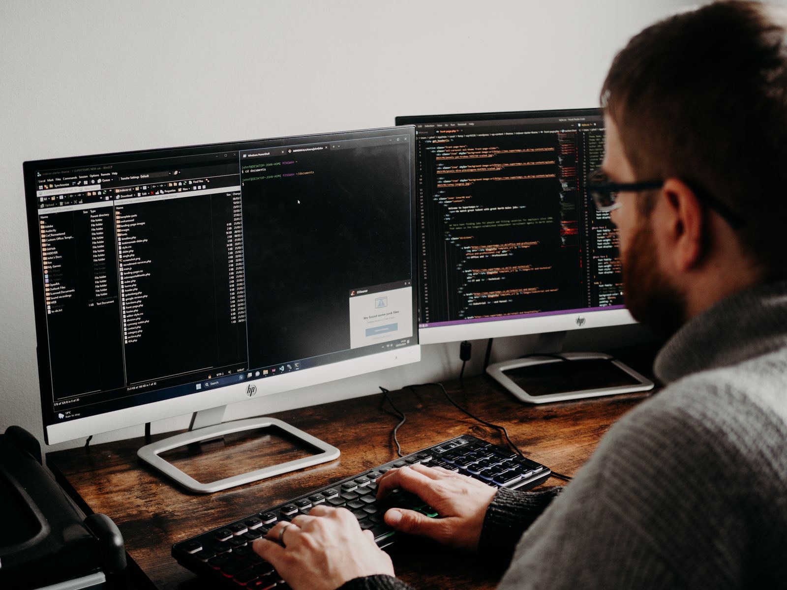 Over-the-shoulder view of a bearded man in glasses and a grey sweater, both hands on a black keyboard at a wooden desk, facing two monitors — the left showing a file manager and a black PowerShell window, the right showing multicoloured source code in a dark editor
