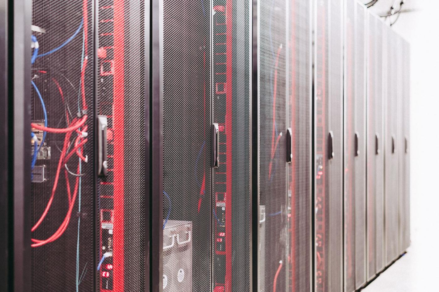 Dãy tủ rack máy chủ chụp chéo dọc hành lang phòng máy tường trắng, tủ gần nhất mở lộ bó dây mạng đỏ và xanh dương chằng chịt cùng vài đèn LED đỏ nhỏ, các tủ phía sau cửa lưới đục lỗ mờ dần về cuối hành lang
