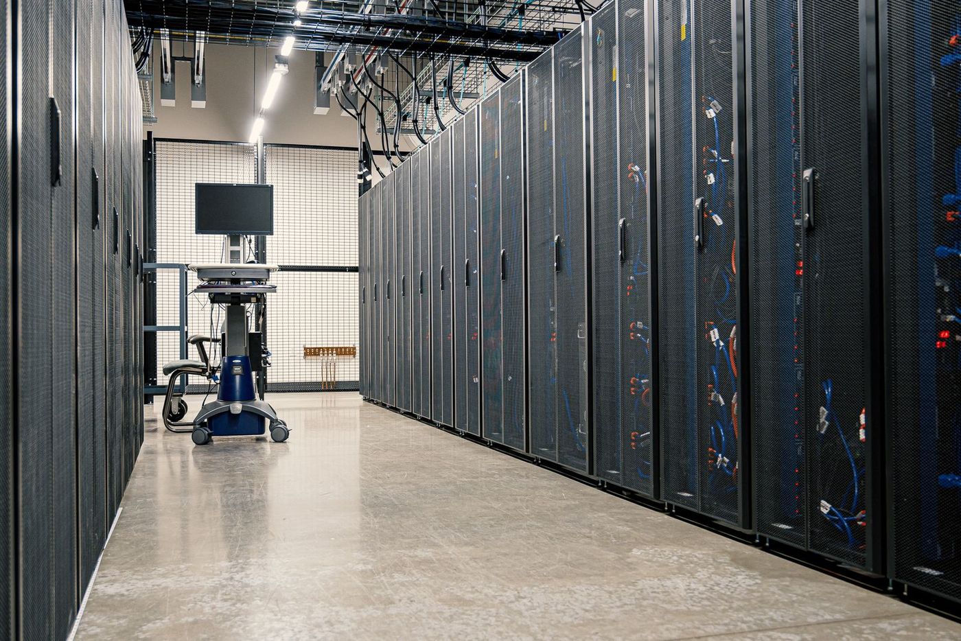 Hành lang trong trung tâm dữ liệu với hai dãy tủ rack lưới đen, cáp mạng xanh và đỏ chạy bên trong, xe đẩy màn hình kỹ thuật ở cuối hành lang