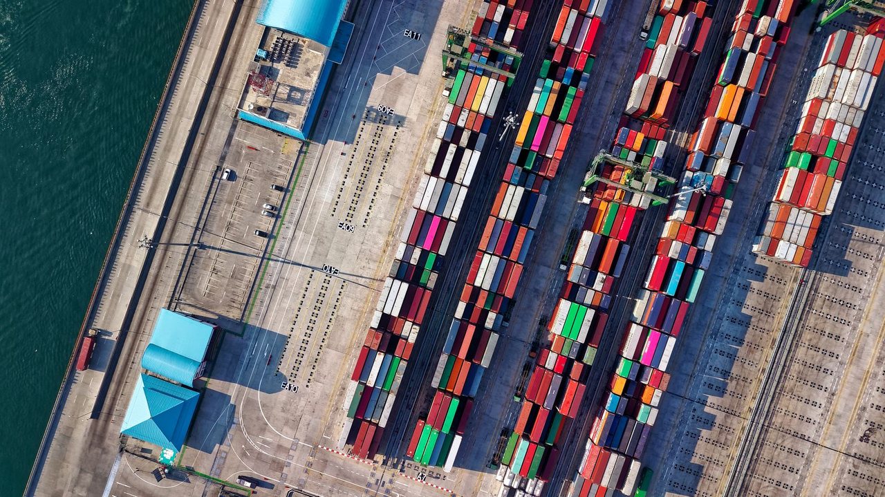Ảnh chụp từ trên cao một cảng container: hàng nghìn container xếp thành các lô song song, bến nước bên trái, cần cẩu xếp dỡ hoạt động