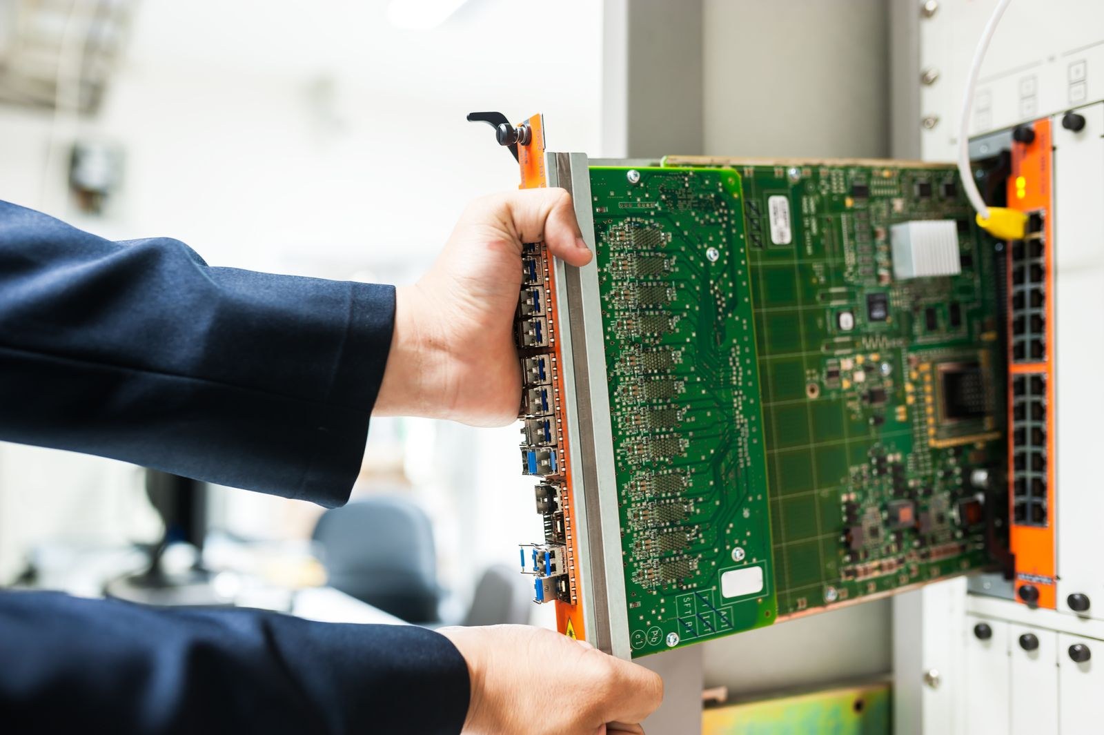 Cận cảnh hai bàn tay trong áo vest xanh navy đang rút một line card mạch xanh lá đầy chip và cổng SFP ra khỏi khung thiết bị mạng trong phòng máy chủ